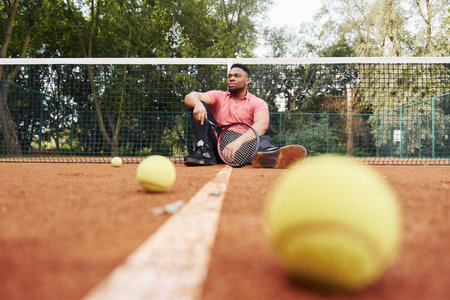 Sits near net and taking a break. African american man in pink shirt sits with tennis racket on the court outdoorsの写真素材