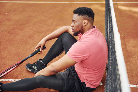 Takes a break. African american man in pink shirt plays tennis on the court outdoorsの写真素材