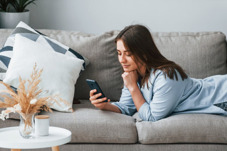 Beautiful young woman in jeans and blue shirt lying down at sofa with phone in handの写真素材