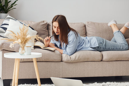 Beautiful young woman in jeans and blue shirt lying down at sofa with notepad and penの写真素材