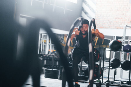 Strong african american man in sportive clothes have workout day in the gym. Doing crossfit by using ropesの写真素材