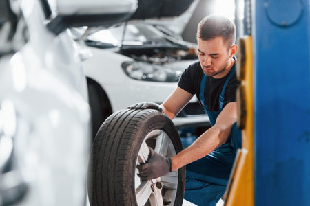 Man in work uniform sitting and changing car wheel indoors. Conception of automobile serviceの写真素材