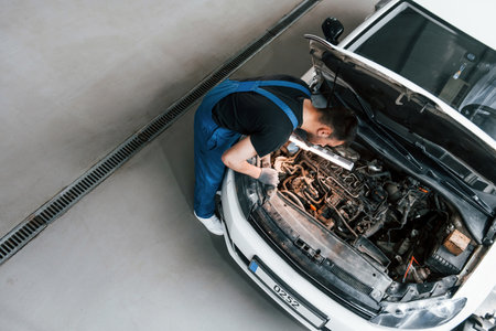 Under the hood. Man in work uniform repairs white automobile indoors. Conception of automobile serviceの写真素材