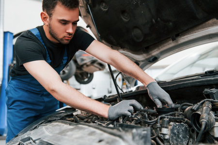 Under the hood. Man in work uniform repairs white automobile indoors. Conception of automobile serviceの写真素材