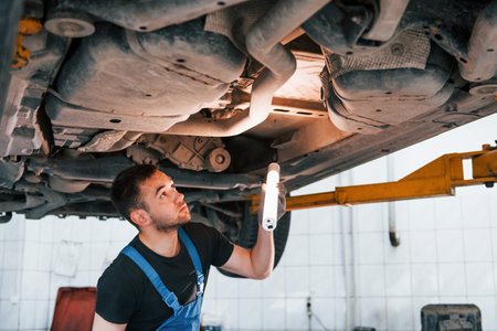Uses lighting equipment. Man in work uniform repairs white automobile indoors. Conception of automobile serviceの写真素材