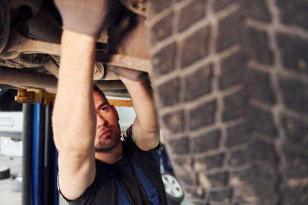 Man in work uniform standing under car and repairs it indoors. Conception of automobile serviceの写真素材