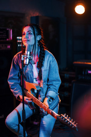 Young beautiful female performer with guitar singing and rehearsing in a recording studioの写真素材