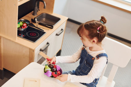 Little girl in casual clothes sits by table and having fun by playing with toys on the kitchenの写真素材