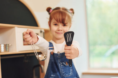 Holding tools in hands and shows it. Little girl in casual clothes have fun by playing with toys on the kitchenの写真素材