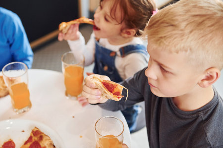 Three children sitting indoors by the table and eating pizza with orange juice togetherの写真素材
