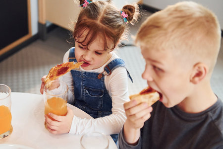 Three children sitting indoors by the table and eating pizza with orange juice togetherの写真素材
