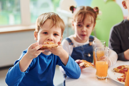 Three children sitting indoors by the table and eating pizza with orange juice togetherの写真素材