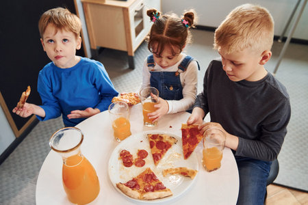 Three children sitting indoors by the table and eating pizza with orange juice togetherの写真素材
