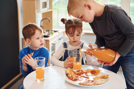 Three children sitting indoors by the table and eating pizza with orange juice togetherの写真素材
