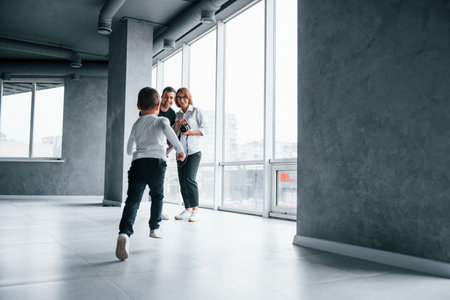 Woman in formal clothes and with camera in hand standing inside of empty room with man and little boy that running and having funの写真素材