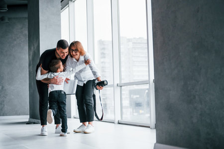 Woman in formal clothes and with camera in hand standing inside of empty room with man and little boy that running and having funの写真素材