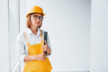 With notepad in hands. Female worker or engineer in yellow uniform and hard hat standing indoorsの写真素材