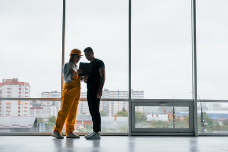 Female worker or engineer in yellow uniform and hard hat standing indoors with manの写真素材