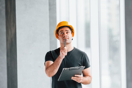 Male worker or engineer in yellow hard hat standing indoors with notepad