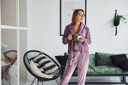 Pregnant young woman in pajamas and glasses standing indoors at daytime