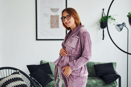 Pregnant young woman in pajamas and glasses standing indoors at daytimeの写真素材