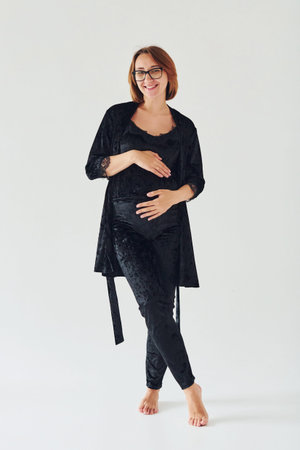 Pregnant young woman in pajamas and glasses standing indoors at daytime against white backgroundの写真素材