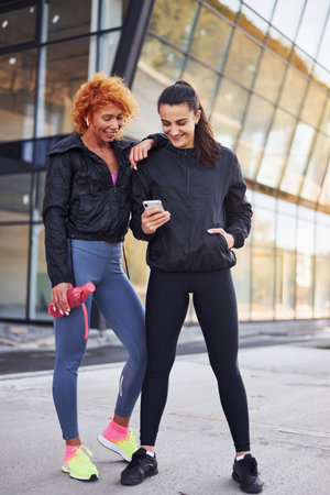 Holds phone in hands. Two female friends with sportive bodies have fitness day outdoors