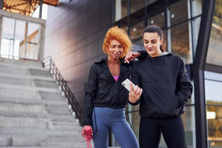 Holds phone in hands. Two female friends with sportive bodies have fitness day outdoorsの写真素材