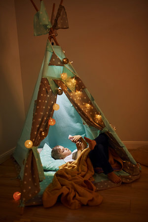 Using phone. Young boy in casual clothes lying down in tent at evening time