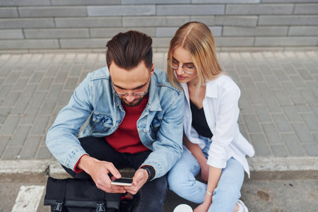 Using phone. Young stylish man with woman in casual clothes sitting outdoors together. Conception of friendship or relationshipsの写真素材