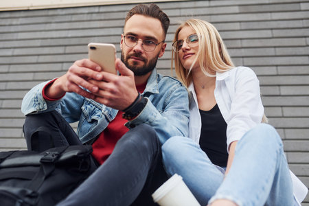 Using phone. Young stylish man with woman in casual clothes sitting outdoors together. Conception of friendship or relationshipsの写真素材