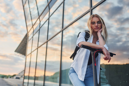 Against modern business building. Beautiful blonde in casual clothes riding electric schooter outdoors at sunny daytimeの写真素材