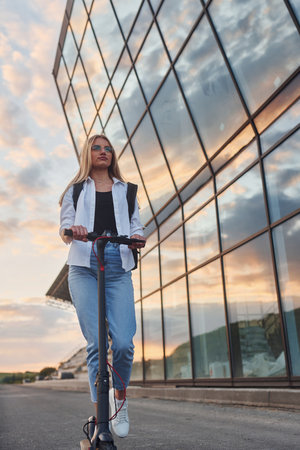 Against modern business building. Beautiful blonde in casual clothes riding electric schooter outdoors at sunny daytimeの写真素材