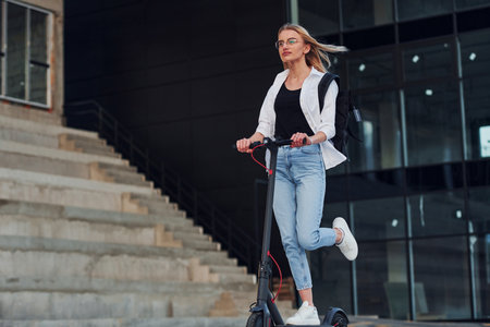 Near business building. Beautiful blonde in casual clothes riding electric schooter outdoors at sunny daytimeの写真素材