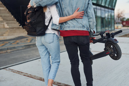 With electric schooter. Young stylish man with woman in casual clothes outdoors near business building together. Conception of friendship or relationshipsの写真素材