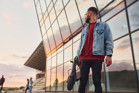 Handsome young guy in casual clothes standing with electric schooter outdoors at sunny daytime near business buildingの写真素材