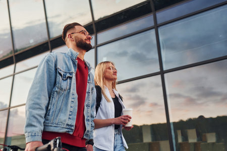 With electric schooter. Young stylish man with woman in casual clothes outdoors near business building together. Conception of friendship or relationshipsの写真素材
