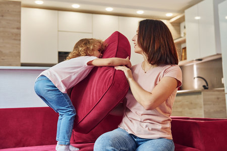 Having fun and jumping. Young mother with her little daughter in casual clothes together indoors at homeの写真素材