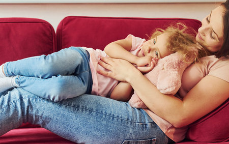 Lying down on red sofa. Young mother with her little daughter in casual clothes together indoors at...