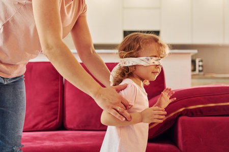 Playing game with eyes blindfolded. Young mother with her little daughter in casual clothes together indoors at homeの写真素材