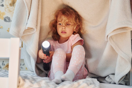 Sitting under blanket with flashlight. Cute little girl in casual clothes is indoors at home at daytimeの写真素材