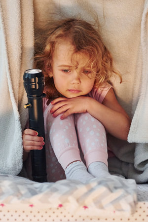 Sitting under blanket with flashlight. Cute little girl in casual clothes is indoors at home at daytimeの写真素材