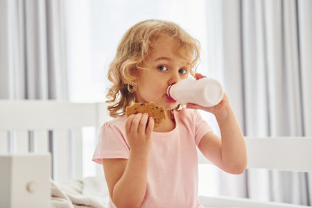 Sitting on the bed, eating cookies and drinking milk. Cute little girl in casual clothes is indoors at home at daytimeの写真素材