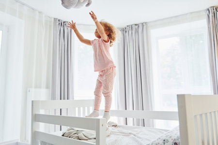 Playing with pillow by throwing it. Cute little girl in casual clothes is indoors at home at daytimeの写真素材