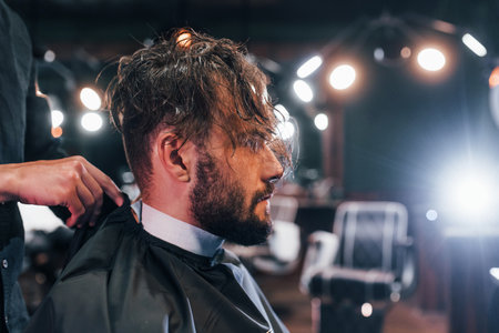 Young bearded man sitting and getting haircut in barber shopの写真素材