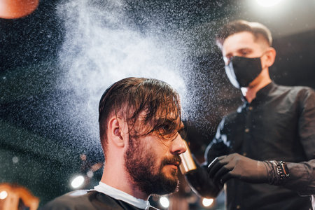 Young bearded man sitting and getting haircut in barber shop by guy in black protective mask
