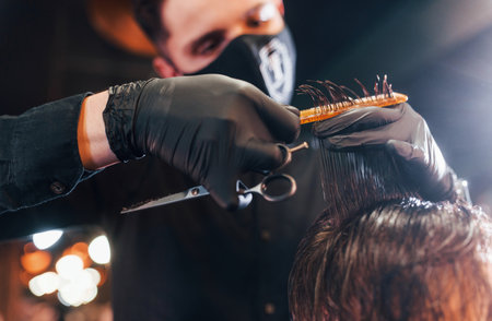 Young bearded man sitting and getting haircut in barber shop by guy in black protective maskの写真素材