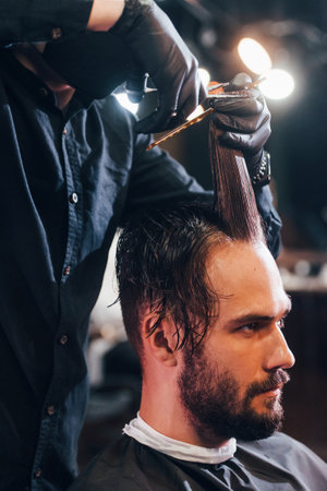 Young bearded man sitting and getting haircut in barber shop by guy in black protective maskの写真素材