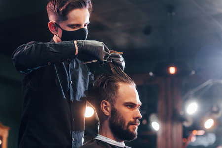 Young bearded man sitting and getting haircut in barber shop by guy in black protective maskの写真素材
