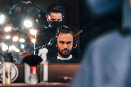 Front view of young bearded man that sitting and getting haircut in barber shop by guy in black protective maskの写真素材